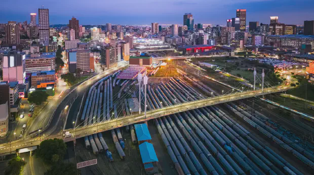 Aerial view at dusk of the Nelson Mandela Bridge crossing a rail yard in Johannesburg, where dozens of tracks fan out beneath the city skyline